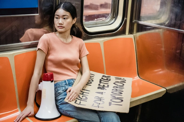 a woman sitting on a train holding a sign, by William Berra, trending on unsplash, feminist art, young asian woman, wearing an orange t shirt, new york in the future, milk bar magazine