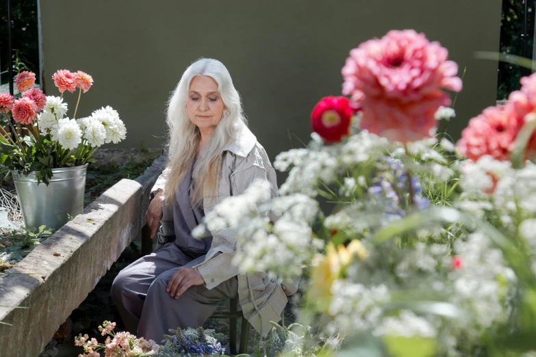 a woman sitting on a bench surrounded by flowers, inspired by Grethe Jürgens, renaissance, light gray long hair, press shot, grey, environmental shot