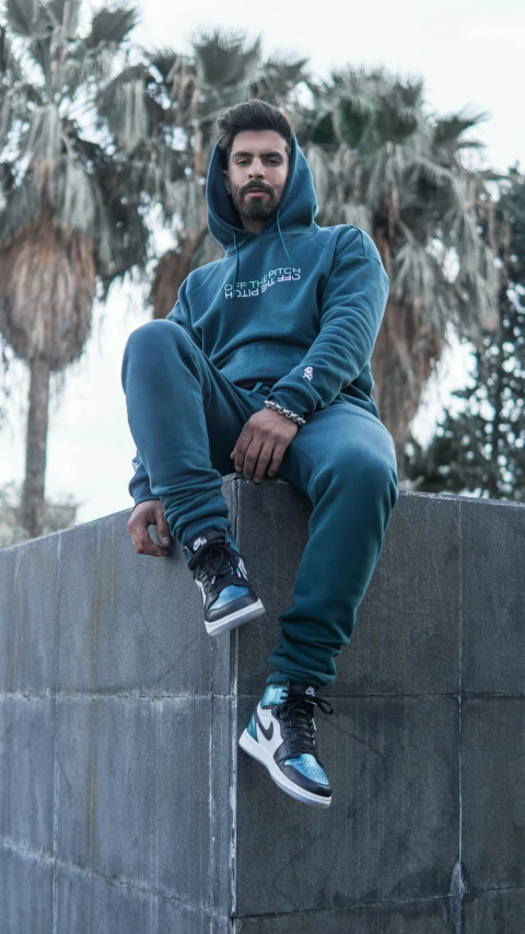 a man sitting on top of a cement wall, an album cover, inspired by Paul Georges, unsplash contest winner, hurufiyya, teal uniform, wearing a hoodie and sweatpants, ( ( dark green, full body 8k