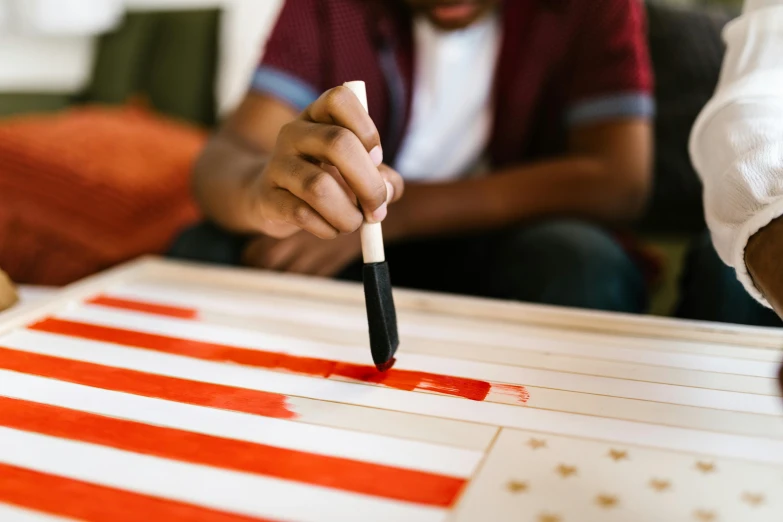 a man is painting an american flag on a table, a child's drawing, by Lee Loughridge, pexels contest winner, red and orange colored, holding a paintbrush, shodo, red and white stripes