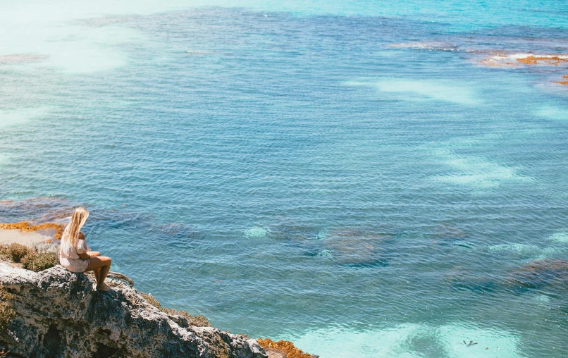 a woman sitting on top of a cliff next to the ocean, by Carey Morris, pexels contest winner, carribean turquoise water, zoomed in, panoramic, colour corrected