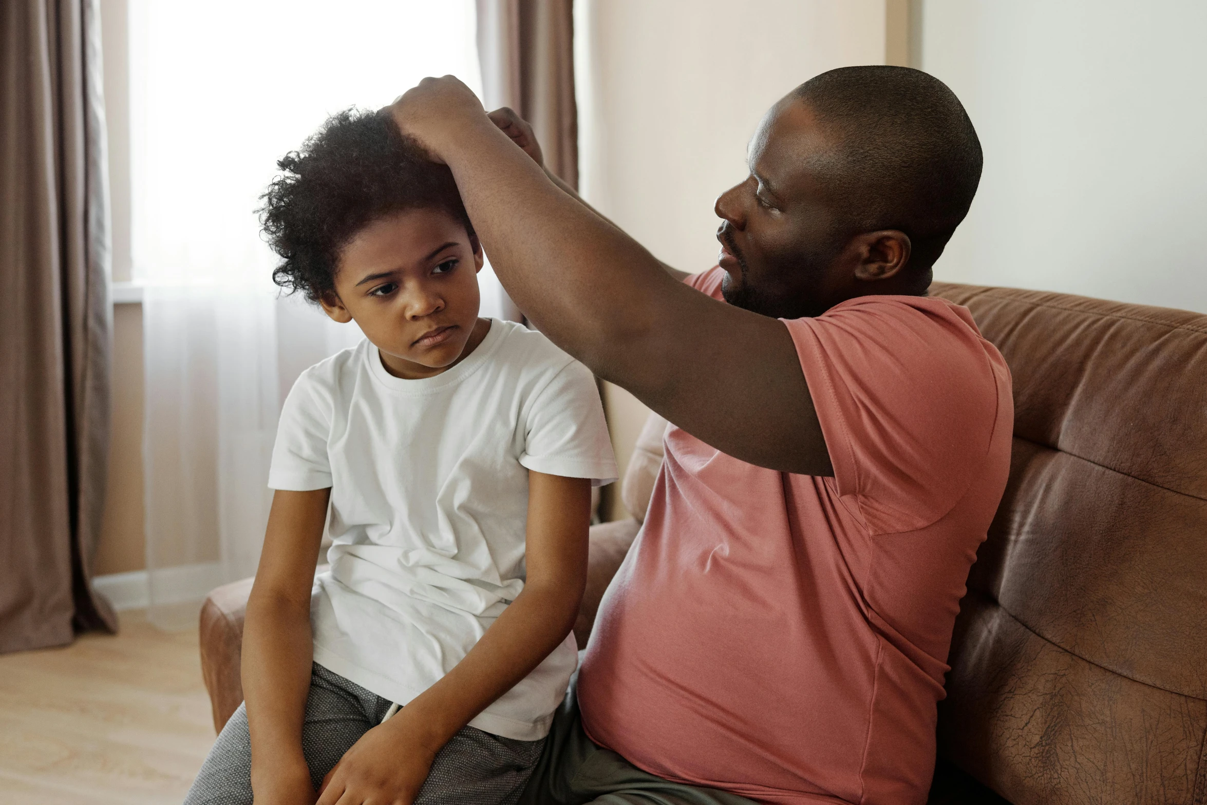 a man combing a young boy's hair while sitting on a couch, by Everett Warner, pexels, hurufiyya, man is with black skin, touching heads, 15081959 21121991 01012000 4k, raising an arm