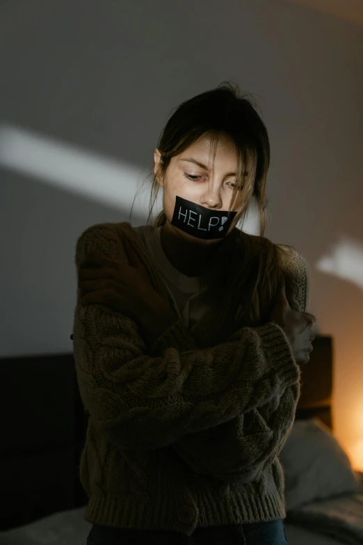 a woman covering her mouth while standing in front of a bed, inspired by Elsa Bleda, trending on pexels, graffiti, black bandage on arms, woman holding sign, wearing a dark sweater, help