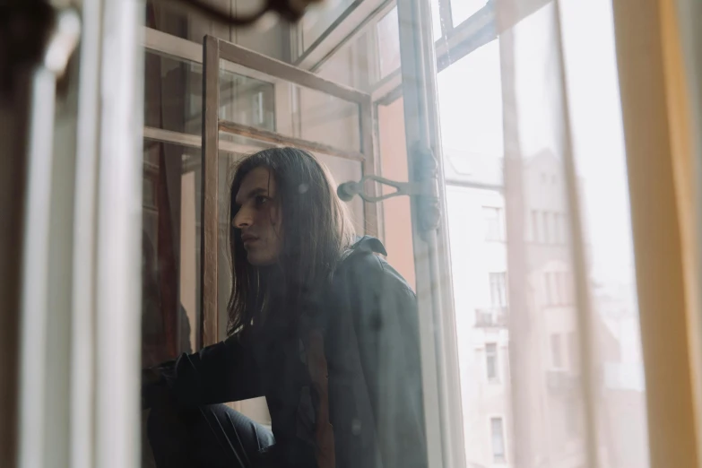 a man sitting on a window sill looking out a window, antipodeans, with long hair, dasha taran, hazy, hammershøi