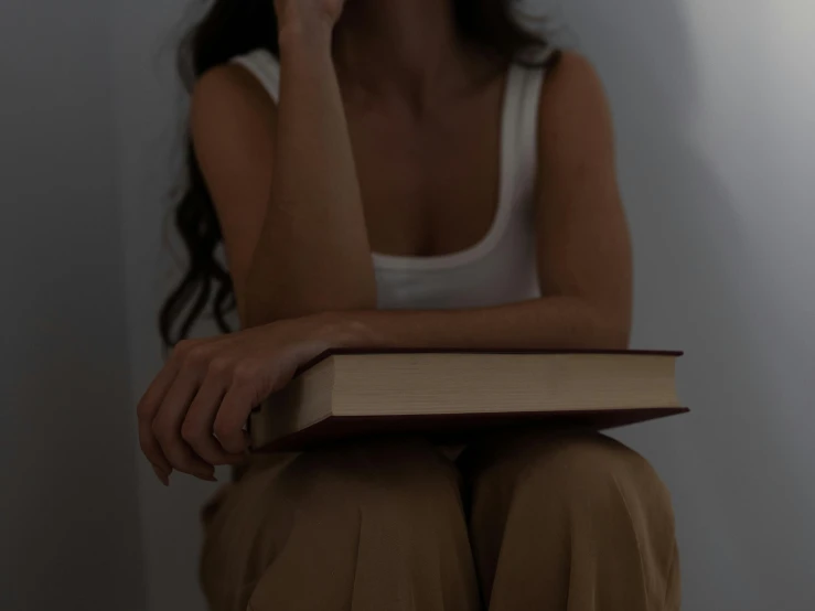 a woman sitting on a chair holding a book, trending on pexels, late night melancholic photo, background image, slightly tanned, curled up on a book