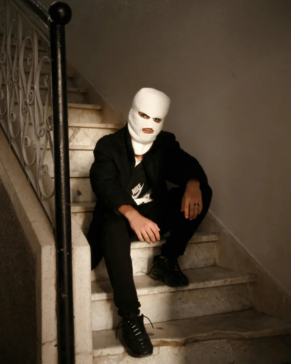 a man in a white mask sitting on a set of stairs, an album cover, unsplash, antipodeans, non-binary, taken in the late 2010s, mina petrovic, the third… had a face like a man