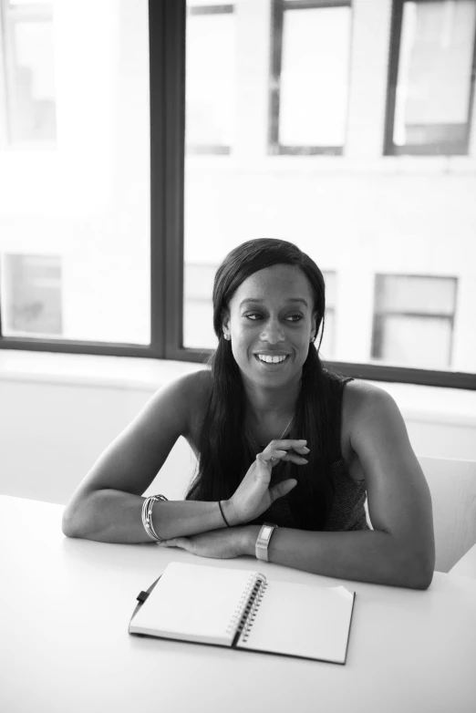 a woman sitting at a table in front of a window, a black and white photo, by Lily Delissa Joseph, ceo, dark skinned, sitting on a desk, darren bartley