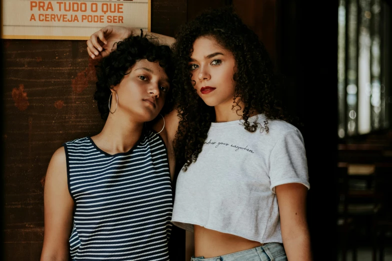 two women standing next to each other in front of a sign, by Elsa Bleda, trending on pexels, antipodeans, curly hair, wearing a t-shirt, brazilian, background image