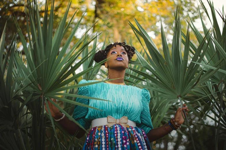 a woman standing in front of a palm tree, by Lily Delissa Joseph, pexels contest winner, afrofuturism, foliage clothing, teal tones, in savannah, cactus and pearls over the head