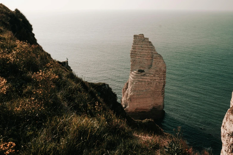 a person standing on top of a cliff next to the ocean, by Raphaël Collin, pexels contest winner, normandy, creating a thin monolith, thumbnail, brown