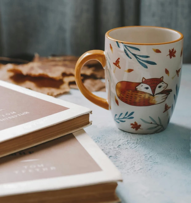 a book sitting on top of a table next to a coffee cup, a still life, trending on pexels, arts and crafts movement, whimsical fox, autumnal, white mug, product introduction photo