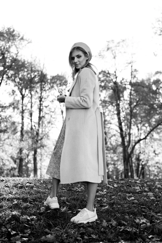 a black and white photo of a woman in a park, wearing a long beige trench coat, autumnal, pastel clothing, promo image