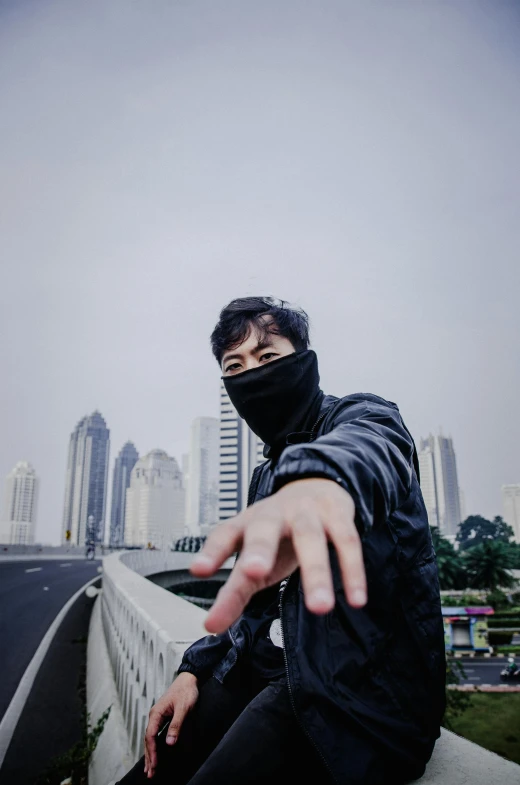 a person sitting on a ledge with a city in the background, an album cover, inspired by Zhu Da, unsplash, realism, balaclava covering face, pointing at the camera, jakarta, headshot profile picture