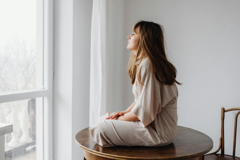 a woman sitting on top of a wooden table next to a window, by Emma Andijewska, trending on pexels, wearing pajamas, girl with brown hair, on a pedestal, calm serene relaxed