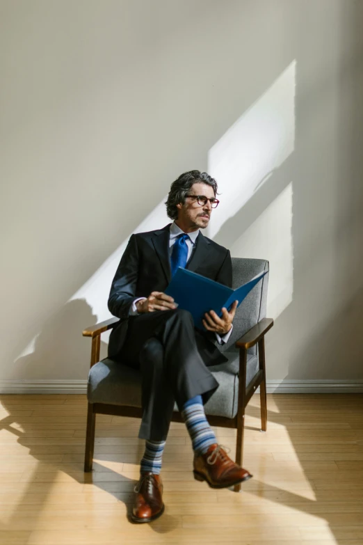 a man sitting in a chair reading a book, wearing a suit and glasses, ben mauro, day light, ignant