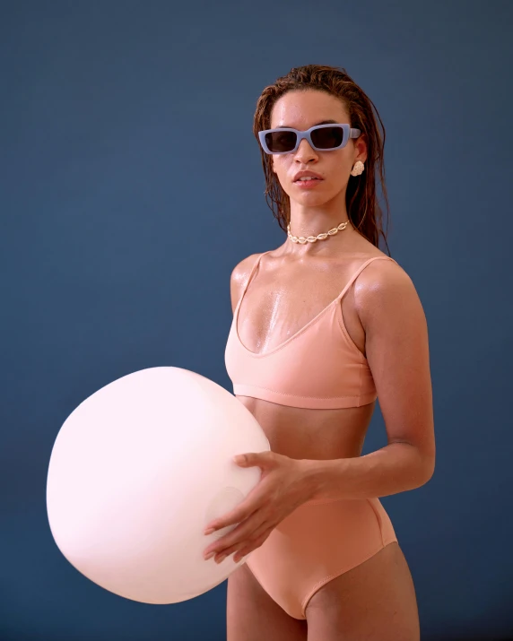 a woman in a bikini holding a white ball, an album cover, inspired by Elsa Bleda, trending on unsplash, bauhaus, fashion model in sunglasses, inflatable, pastel pink concrete, portrait mode photo