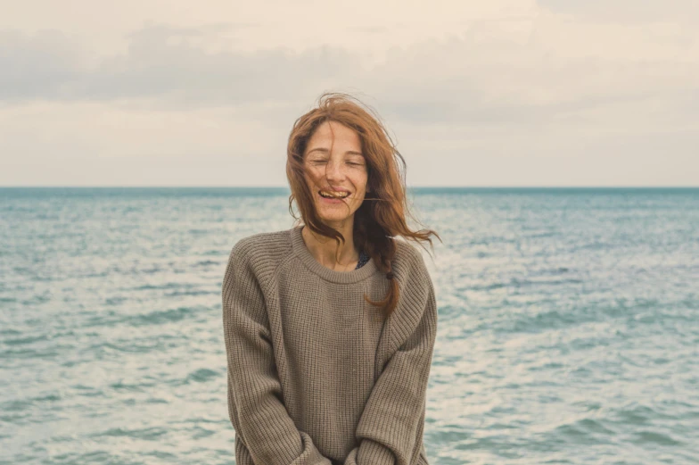 a woman standing in front of a body of water, pexels contest winner, sarcastic smiling, brown sweater, avatar image, at the seaside