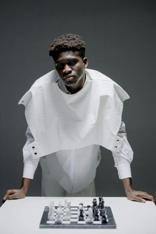 a man sitting at a table with a chess board, inspired by Paul Georges, afrofuturism, ripped up white garment, model standing pose, with shoulder pads, showstudio