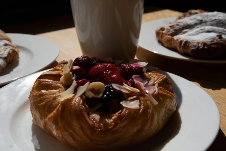 a pastry sitting on top of a white plate next to a cup of coffee, inspired by Richmond Barthé, unsplash, renaissance, plates of fruit, in a sunbeam, chilean, raspberry