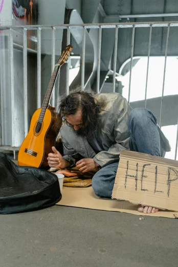 a man sitting on the ground with a guitar, by Francis Helps, homeless, soup, promo image, getty images