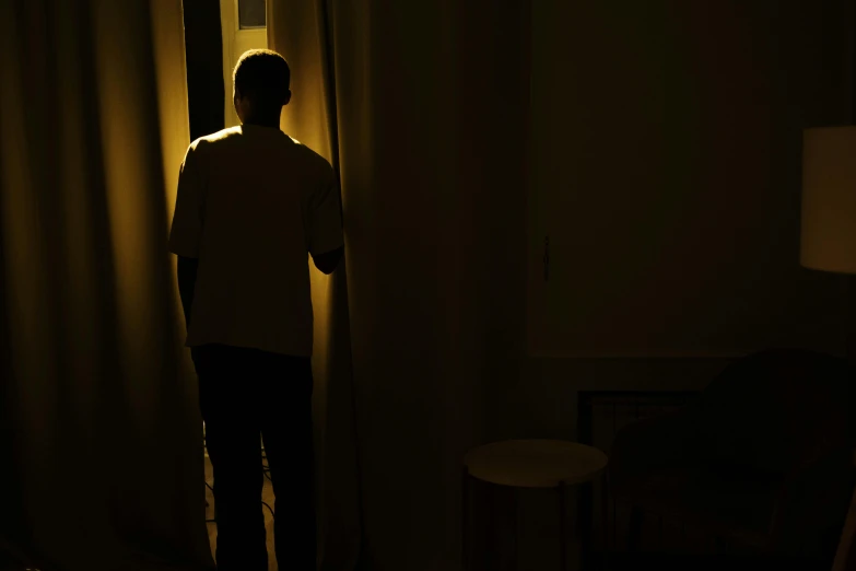 a man standing in a dimly lit room, happening, looking out the window, no lights in bedroom, yellow light, glowing drapes