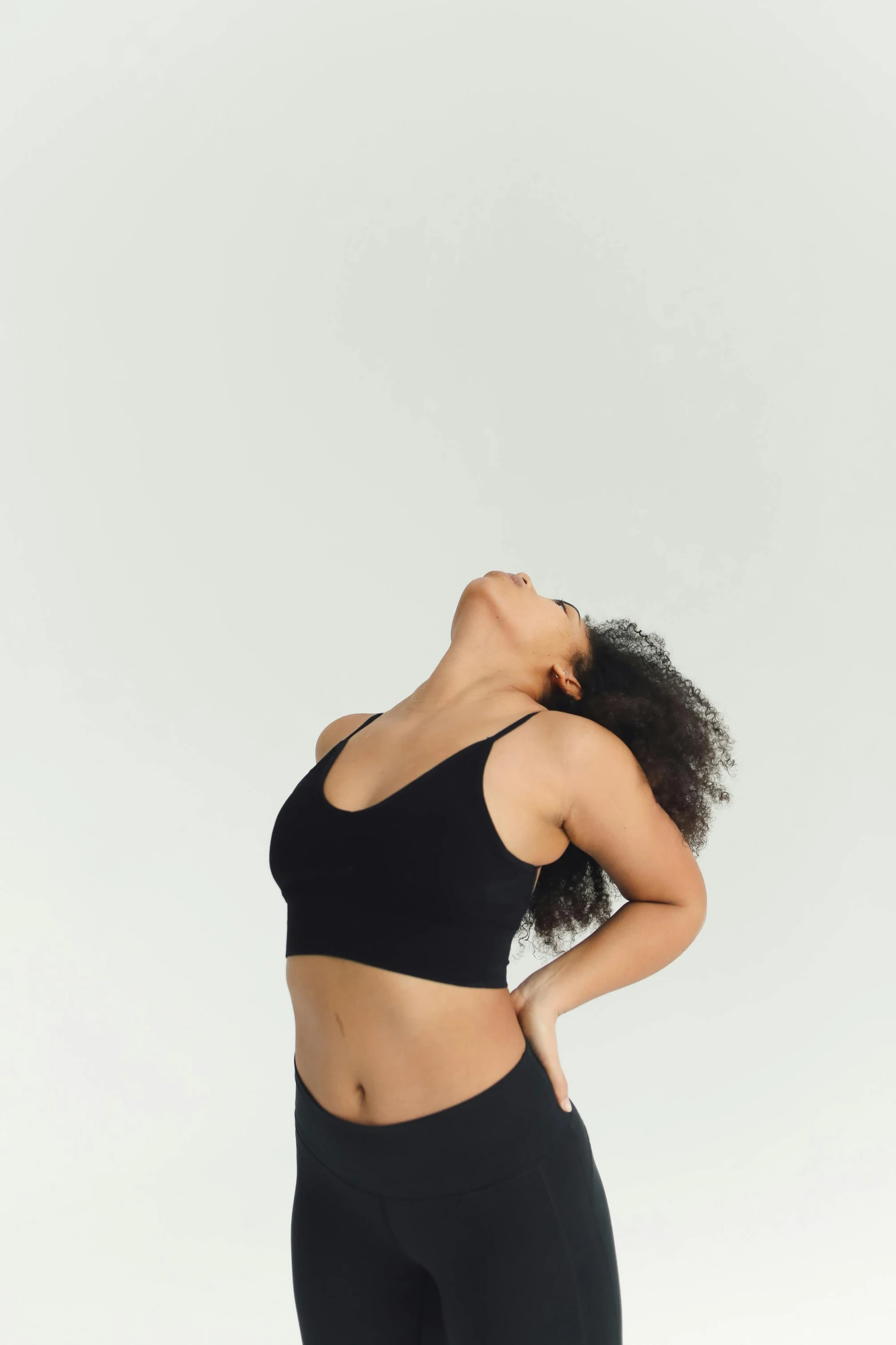 a woman in a black sports bra top and black leggings, by Carey Morris, trending on unsplash, renaissance, meditative sacral pose, in front of white back drop, young woman looking up, woman with black hair