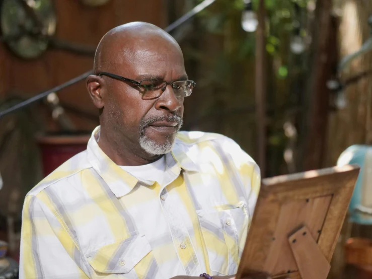 a man sitting in front of a painting on a easel, an album cover, inspired by George Barret, Sr., pexels contest winner, hurufiyya, wearing reading glasses, still image from tv series, emmanuel shiru, gardening