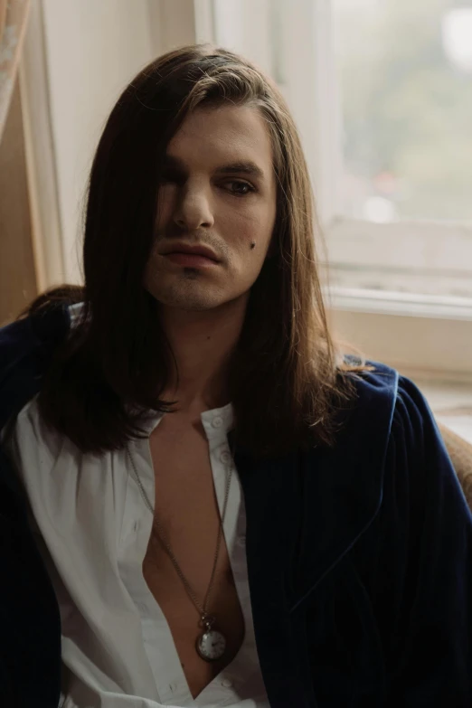 a man sitting on top of a couch next to a window, an album cover, inspired by Nan Goldin, renaissance, with long hair and piercing eyes, delicate androgynous prince, andrea savchenko, wearing cross on robe