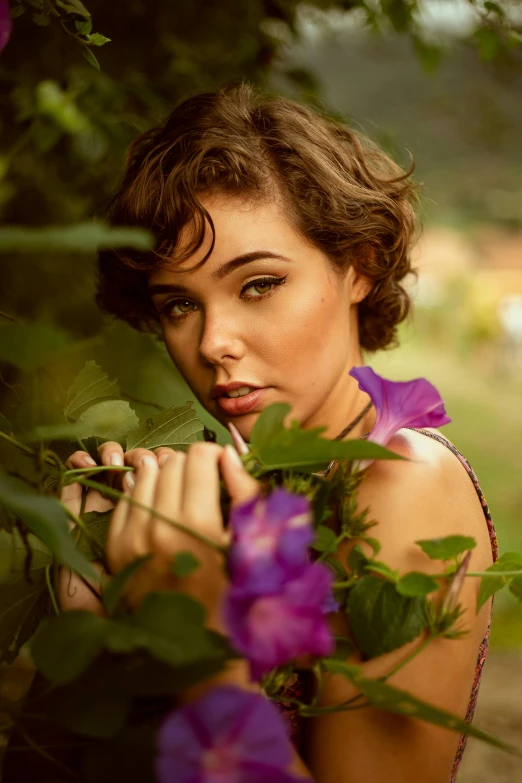 a woman standing next to a bush with purple flowers, a colorized photo, by Galen Dara, pexels contest winner, curly pixie cut hair, brazilan supermodel, attractive young woman, alluring eyes