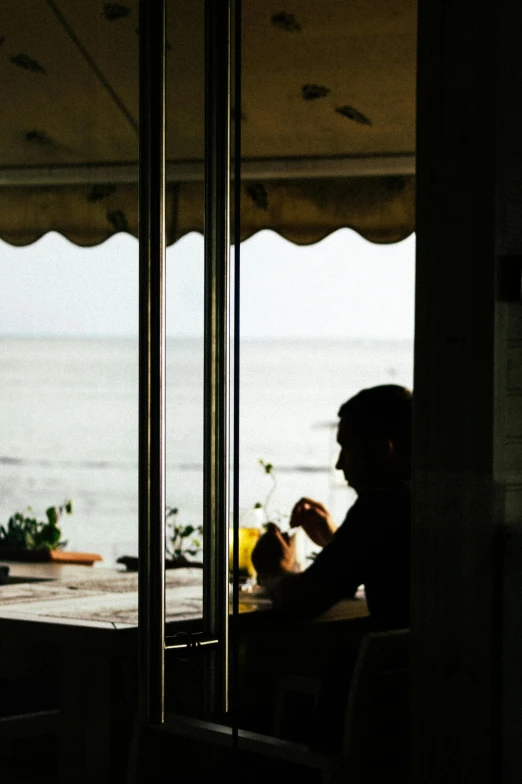 a man sitting at a table in front of a window, a picture, unsplash, beach bar, silhouette :7, eating, film photo