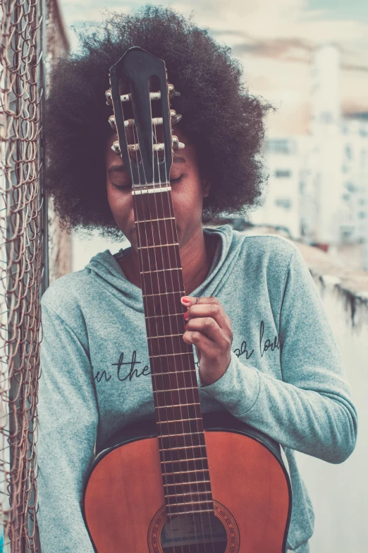 a woman holding a guitar next to a tree, by Matija Jama, pexels contest winner, happening, brown skinned, wearing a grey hooded sweatshirt, afro, instagram post