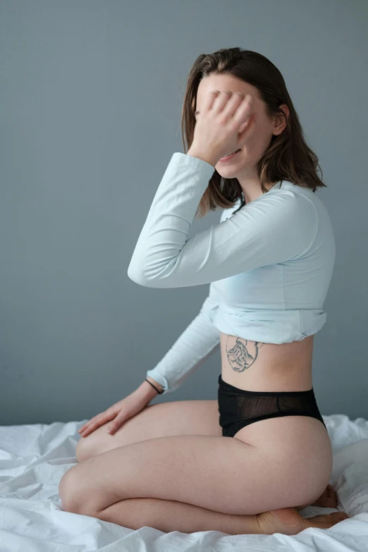 a woman sitting on a bed covering her face, a tattoo, her belly button is exposed, pale blue skin, plain background, trending photo