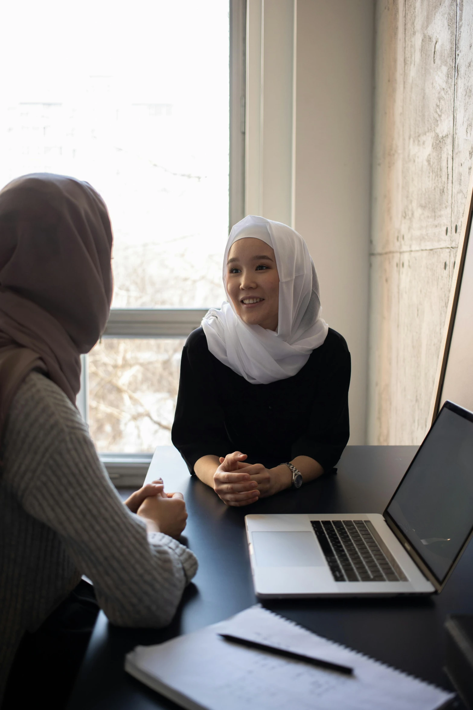 two women sitting at a table talking to each other, hurufiyya, using a macbook, wearing a head scarf, asian human, sitting in dean's office