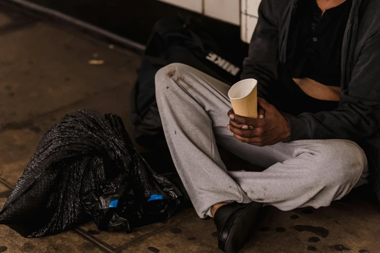 a man sitting on the ground holding a cup, a photo, trending on unsplash, excessivism, mad trash bags, in a subway, black man, malnourished