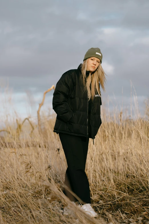 a woman standing in a field of tall grass, an album cover, inspired by Louisa Matthíasdóttir, unsplash, model wears a puffer jacket, black beanie, outlive streetwear collection, full body profile pose