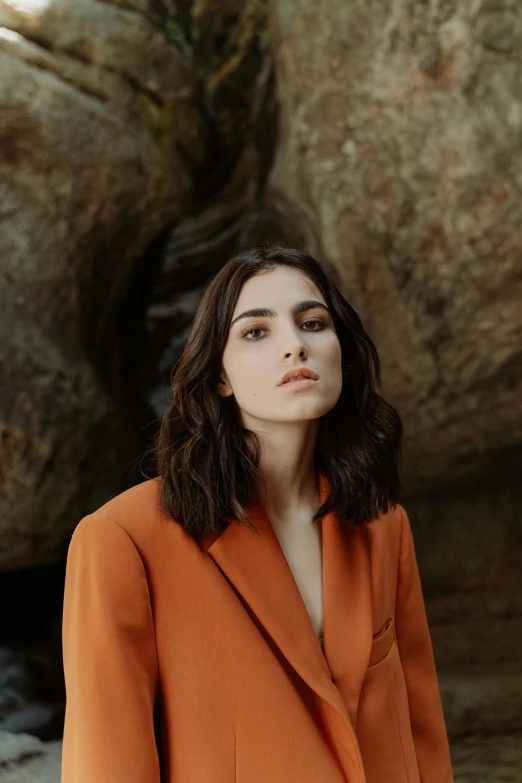 a woman standing in front of a large rock, an album cover, trending on pexels, photorealism, orange jacket, sasha grey, in a suit, hila klein