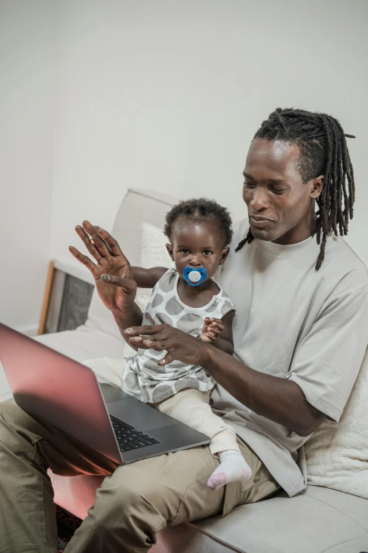 a man sitting on a bed holding a baby and using a laptop, pexels contest winner, figuration libre, papa legba, caring fatherly wide forehead, afro tech, in an action pose