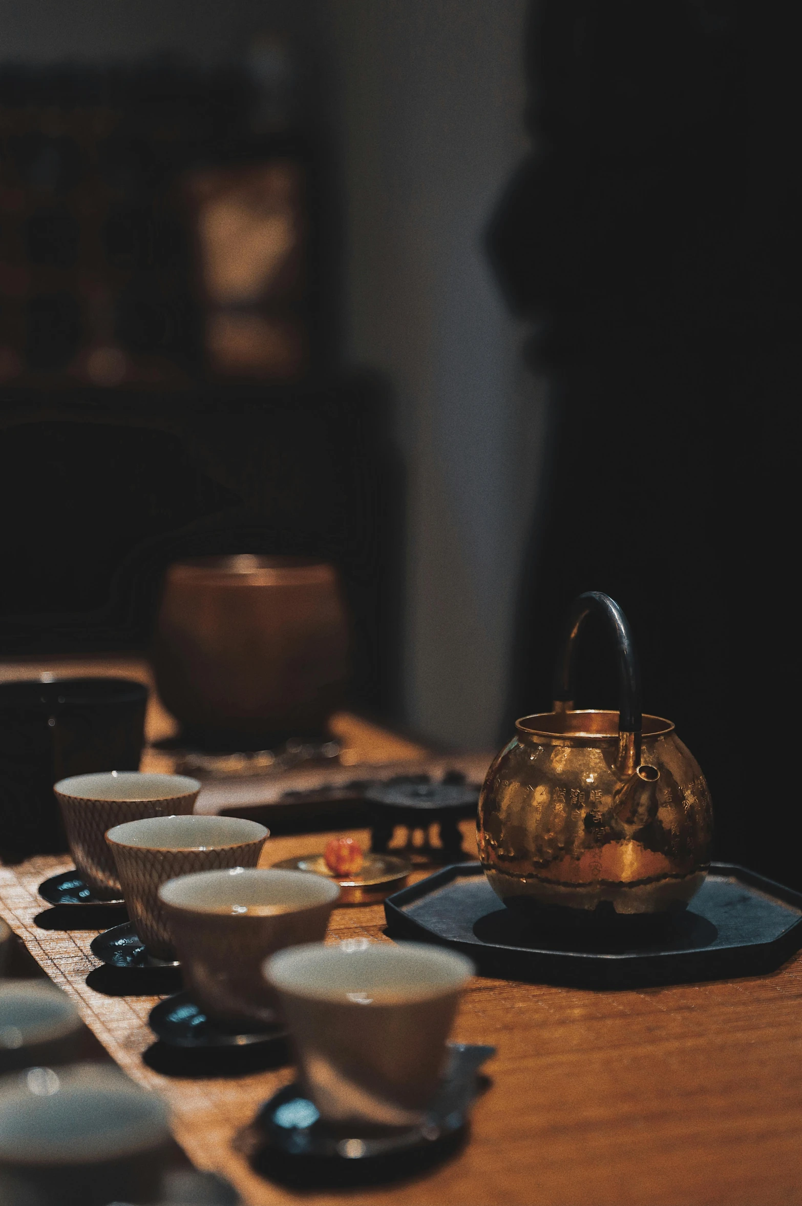 a wooden table topped with cups and saucers, a still life, inspired by Kanō Shōsenin, trending on unsplash, mingei, gold and black color scheme, low light, square, teapot