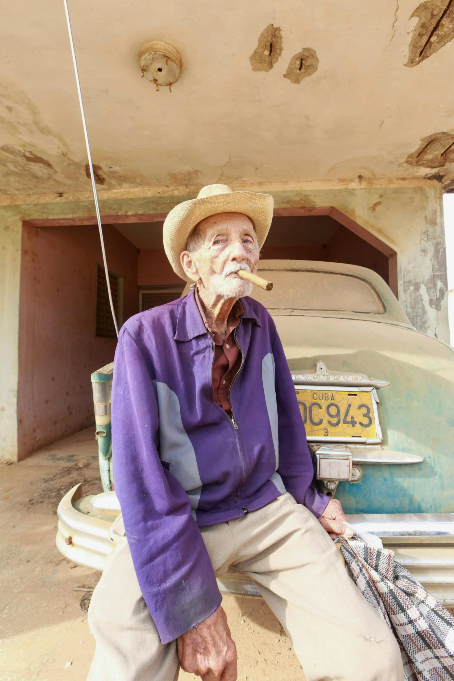 an old man sitting in front of a car, by William Berra, pexels contest winner, cuban setting, color image, tall farmer, with a cigarette in its mouth