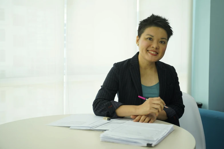 a woman sitting at a table with a pen in her hand, inspired by Ina Wong, corporate photo, slightly smiling, full colour, on a white table