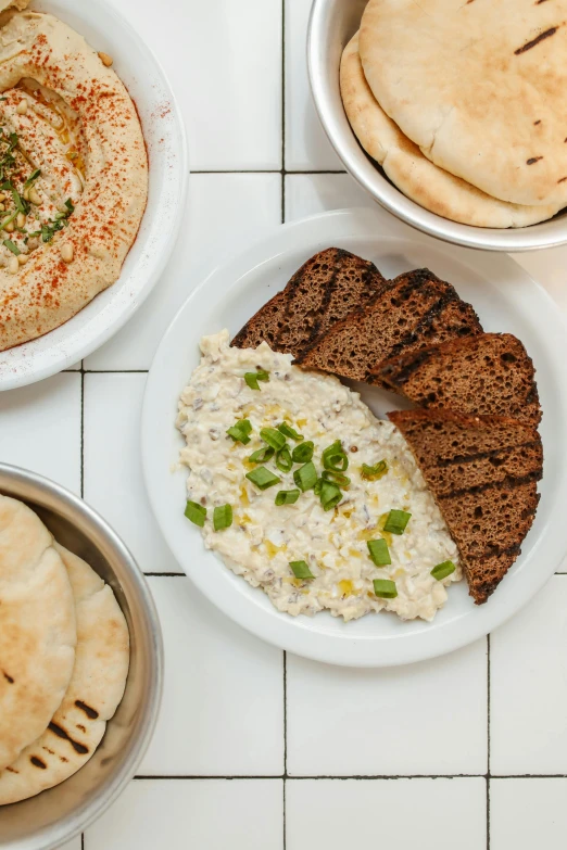 a table topped with plates of food and pita bread, dau-al-set, creamy, speckled, dan mumfor, sleek white