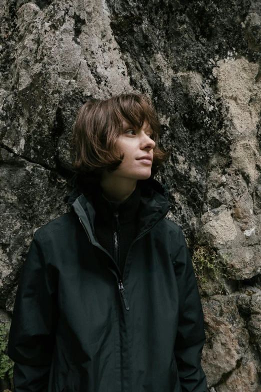 a man standing in front of a rock wall, an album cover, by Attila Meszlenyi, unsplash, girl with brown hair, bob cut, she wears a jacket, botanicals