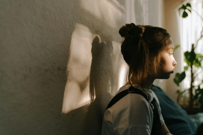 a woman sitting on a couch next to a window, trending on pexels, hyperrealism, hood and shadows covering face, girl with messy bun hairstyle, standing under a beam of light, teenage female schoolgirl