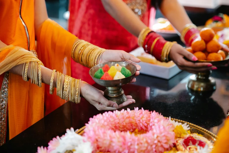 a group of people standing around a table filled with food, hindu ornaments, pink and orange, bangles, holding flowers