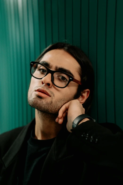 a man wearing glasses leaning against a green wall, an album cover, inspired by Antonio Saura, claudio bravo, looking away from camera, on black background, young spanish man