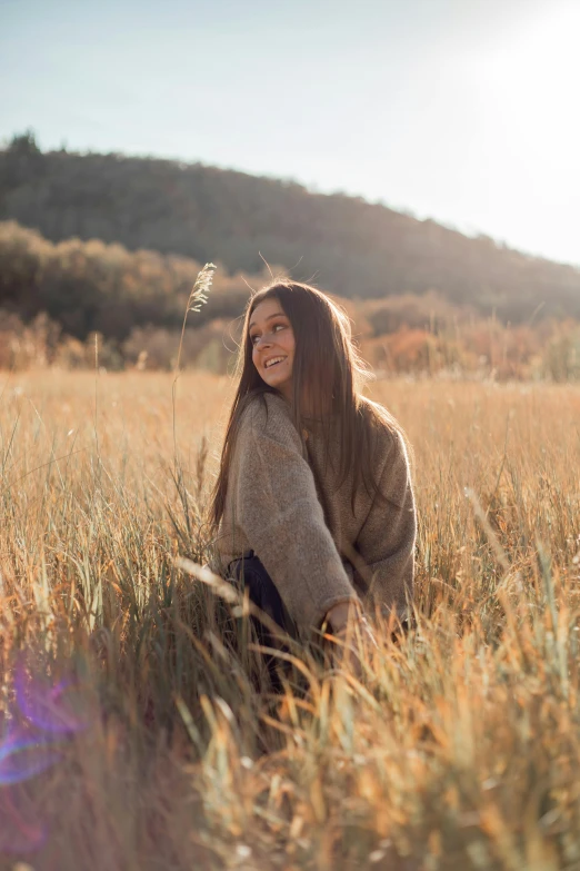 a woman sitting in a field of tall grass, trending on unsplash, 🍂 cute, large smile, brown long and straight hair, wearing an oversized sweater