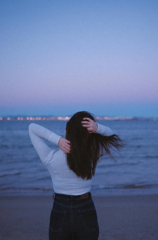 a woman standing on top of a beach next to the ocean, trending on pexels, happening, calm night. over shoulder shot, hands in her hair. side-view, plain background, looming over a city