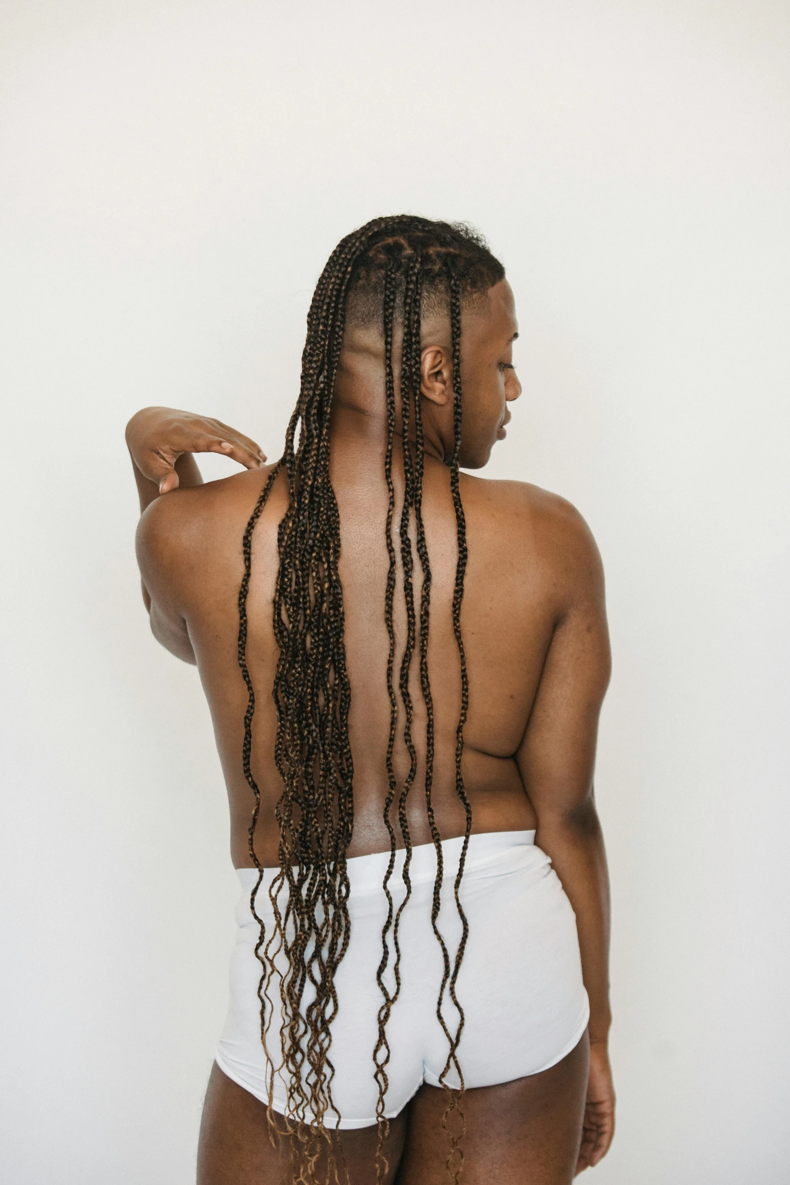 a woman with long hair standing in front of a white wall, an album cover, inspired by Ras Akyem, trending on unsplash, renaissance, his back is turned, cornrows, disrobed, non binary model