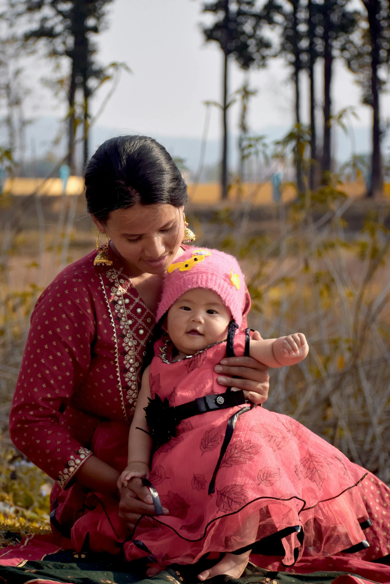 a woman sitting on a blanket holding a baby, background: assam tea garden, pink, medium-shot, hero