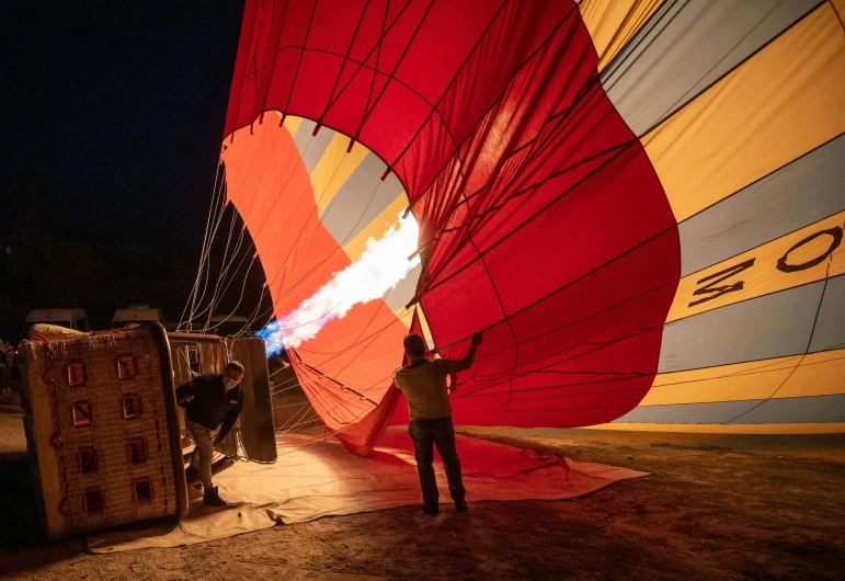 a man standing in front of a hot air balloon, set at night, filling the frame, helmet is off, red and yellow light